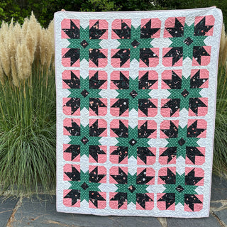 Present Parcels Quilt Pattern by A Piece of Quiet Quilts cover: pink, dark green, and cream geometric star quilt displayed outdoors against ornamental grasses.