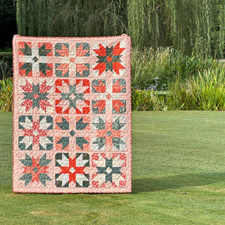 A colorful quilt with red, green, and beige geometric star and parcel blocks displayed outdoors on grass, showcasing the Present Parcels Quilt Pattern (Download) by A Piece of Quiet Quilts.