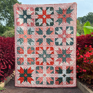 A colorful quilt made with red, green, and pink fabric blocks displayed outdoors among vibrant foliage, showcasing the Present Parcels Quilt Pattern (Download) by A Piece of Quiet Quilts.
