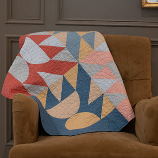 Colorful modern quilt with geometric shapes draped over a brown chair, showcasing the Relative Time Quilt Pattern (Download) by Wood Lily Studio.