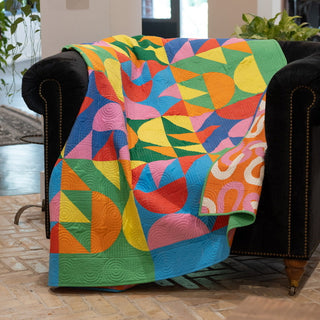 Relative Time Quilt Pattern by Wood Lily Studio lifestyle: colorful geometric quilt with half circles and triangles draped over a black leather armchair in an indoor setting.