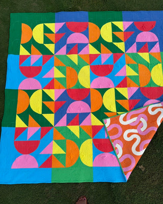 Colorful modern quilt with geometric shapes in bright pink, yellow, orange, green, blue, and red, paired with a contrasting patterned backing, showcasing the Relative Time Quilt Pattern (Download) by Wood Lily Studio.