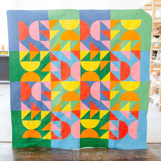 Relative Time Quilt Pattern by Wood Lily Studio cover: bold Bauhaus-style quilt with half circles and triangles in orange, yellow, pink, blue, green, and teal on a wood floor.