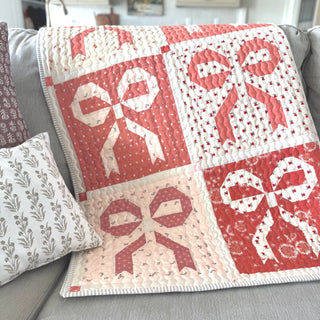 Quilt featuring pink and white bow blocks displayed on a couch, showcasing the Ribbons & Bows Quilt Pattern (Download) by Sew & Sew Quilting Co.