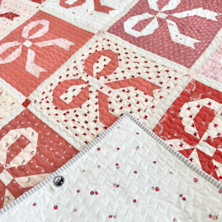 Close-up of a pink and coral fabric quilt with bow and ribbon blocks, featuring a white backing with small strawberry prints, showcasing the Ribbons & Bows Quilt Pattern (Download) by Sew & Sew Quilting Co.