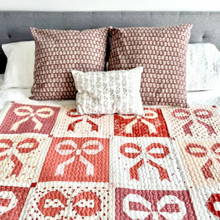 Close-up of a soft quilt featuring ribbon bow designs in warm pink and coral tones, styled on a bed with patterned pillows, showcasing the Ribbons & Bows Quilt Pattern (Download) by Sew & Sew Quilting Co.