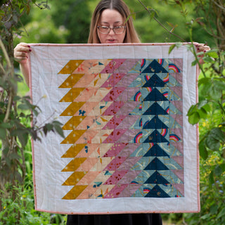 Woman holding a colorful baby quilt made with the Ripple Quilt Pattern (Download) by Lou Orth Designs featuring geometric flying geese blocks in warm and cool tones.