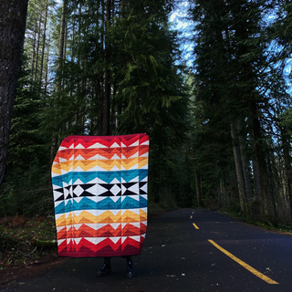 Person standing on a forest road holding a colorful geometric quilt featuring reds, yellows, blues, and blacks, showcasing the Rocky Coast Quilt Pattern (Download) by Toad & Sew.