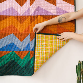 Hands hold a colorful geometric quilt with warm orange, pink, green, and blue tones showing its yellow plaid backing, featuring the Rocky Coast Quilt Pattern (Download) by Toad & Sew.