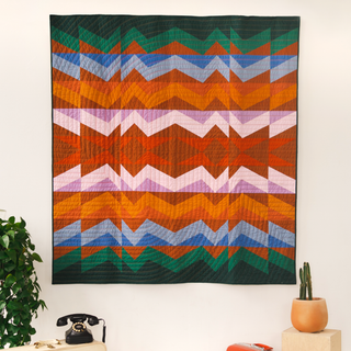 Colorful Rocky Coast quilt by Toad & Sew hanging on a wall with a plant and phone below.