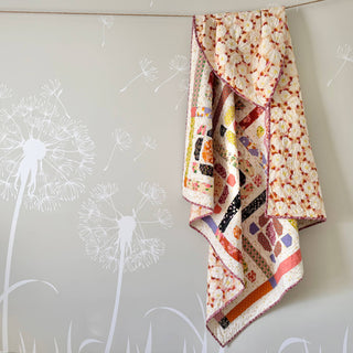 Quilt with colorful geometric and floral prints draped against a soft wall with dandelion decals, showcasing The Dandy Patch’s Saltbox Quilt Pattern (Download).
