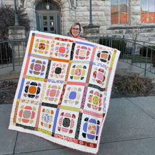 Woman holding a colorful homemade quilt with floral and geometric blocks outside a stone building, showcasing the Saltbox Quilt Pattern (Download) by The Dandy Patch.
