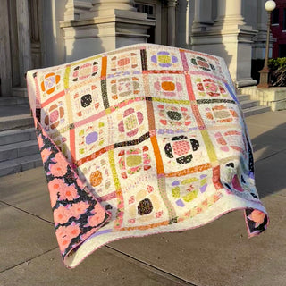 A colorful quilt with floral and geometric blocks displayed outdoors on stone steps, showcasing a delicate mix of pinks, oranges, and purples by The Dandy Patch using the Saltbox Quilt Pattern (Download).