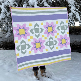 Person holding a pastel colored quilt with star and geometric block patterns in a snowy outdoor setting, showcasing Scatter Sunshine Quilt Pattern (Download) by Shadow Valley Quilts.