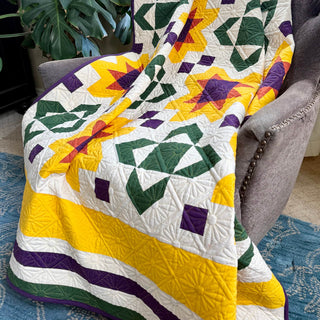 Colorful quilt with purple, green, yellow, and white geometric patterns draped on a chair, showcasing Shadow Valley Quilts' Scatter Sunshine Quilt Pattern (Download).