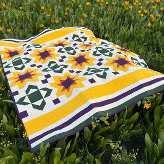 Bright yellow, green, and purple geometric star quilt laid on green grass with yellow flowers, showcasing Scatter Sunshine Quilt Pattern (Download) by Shadow Valley Quilts.