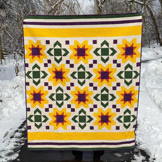 Bright quilt with geometric star and square block patterns in yellow, green, and purple, displayed outdoors in a snowy setting, featuring Scatter Sunshine Quilt Pattern (Download) by Shadow Valley Quilts.