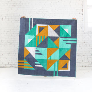 Geometric quilt with teal, mustard, white, and navy blue shapes displayed against a white brick wall, showcasing the Sincerely Quilt Pattern (Download) by Eudaimonia Studio.