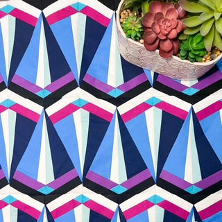 Close-up of a colorful geometric quilt block pattern in blue, purple, pink, and black tones with a succulent planter, featuring the Skye FPP Quilt Pattern (Download) by Julia Wachs Designs.