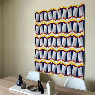 Modern geometric quilt wall hanging in navy, purple, orange, and yellow tones displayed above a dining table with minimalist decor, showcasing Skye FPP Quilt Pattern (Download) by Julia Wachs Designs.