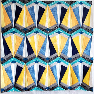 Colorful geometric quilt blocks in blue, yellow, and gray tones arranged in a repeating pattern, showcasing the Skye FPP Quilt Pattern (Download) by Julia Wachs Designs.