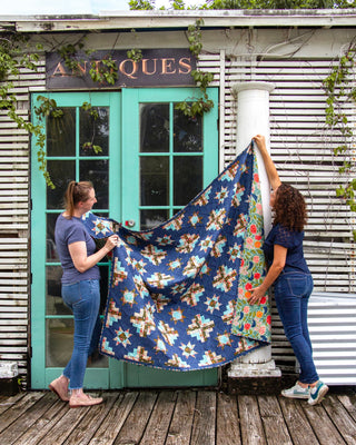 Two women holding up a colorful quilt with starburst and floral patterns outside a rustic building, showcasing the Solstice Burst Quilt Pattern (Download) by A Piece of Quiet Quilts.
