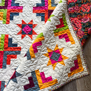 Close-up of a colorful quilt with star and geometric patterns featuring bright reds, pinks, greens, yellows, and blues, showcasing the Solstice Burst Quilt Pattern (Download) by A Piece of Quiet Quilts.
