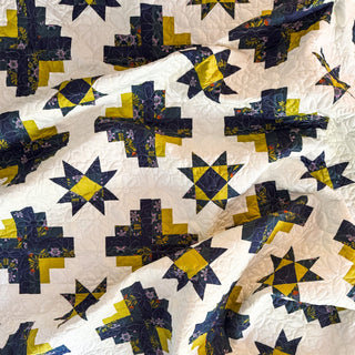 Close-up of a navy and yellow star and cross patterned quilt showcasing detailed stitching, featuring the Solstice Burst Quilt Pattern (Download) by A Piece of Quiet Quilts.