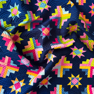 Colorful geometric star and cross quilt blocks in bright pink, yellow, and blue shades on a dark background, showcasing Solstice Burst Quilt Pattern (Download) by A Piece of Quiet Quilts.