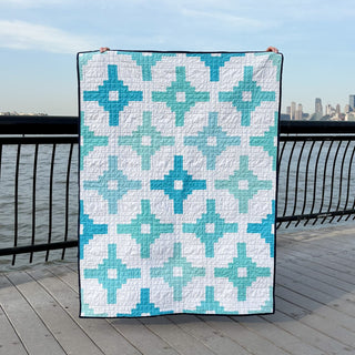 A vibrant quilt featuring a geometric design in various shades of blue and teal displayed outdoors near a waterfront, showcasing the Southwest Knit Quilt Pattern (Download) by Month of May Quilting.