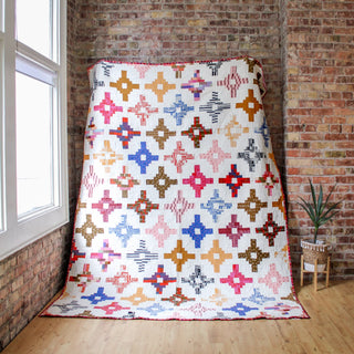 Large colorful quilt with geometric patterns in warm and cool tones displayed against a brick wall, featuring the Southwest Knit Quilt Pattern (Download) by Month of May Quilting.