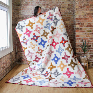Woman holding a large colorful quilt with geometric star-like patterns in warm tones, showcasing the Southwest Knit Quilt Pattern (Download) by Month of May Quilting.