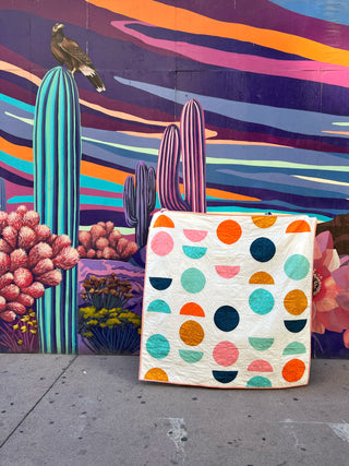 Colorful Spring Moon quilt by Lou Orth Designs in front of a vibrant mural with cacti and flowers.