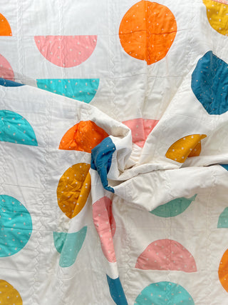 Close-up of a colorful quilt featuring half-circle geometric shapes in orange, pink, teal, blue, and mustard on a white background, designed by Lou Orth Designs using the Spring Moon Quilt Pattern (Download).