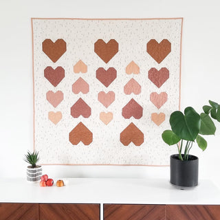Heart-patterned quilt on a white wall with two potted plants below.