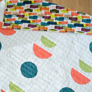 Close-up of a colorful quilt with geometric shapes featuring teal, coral, and lime green on a white background, styled with a multicolored brushstroke patterned backing, showcasing the Sunroom Quilt Pattern (Download) by Sewn.