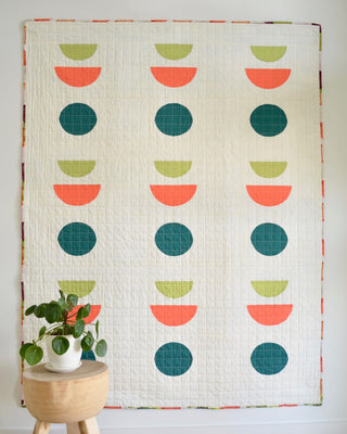 Sunroom Quilt Pattern by Sewn brights colorway on wall: cream quilt with curved half-circle blocks in coral, teal, and olive green, styled with a plant on a wood side table.