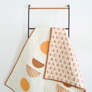 Quilt made with the Sunroom Quilt Pattern (Download) by Sewn, displayed on a black ladder, featuring warm yellow and orange geometric shapes and a soft patterned backing.