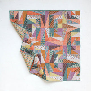 Colorful geometric quilt with various patterned fabrics in warm and cool tones, folded at one corner, showcasing intricate piecing from Eudaimonia Studio’s Super Scrappy Repair Quilt Pattern (Download).