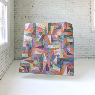 Colorful geometric scrap quilt displayed against a white brick wall, showcasing intricate piecing and pattern design by Eudaimonia Studio using the Super Scrappy Repair Quilt Pattern (Download).