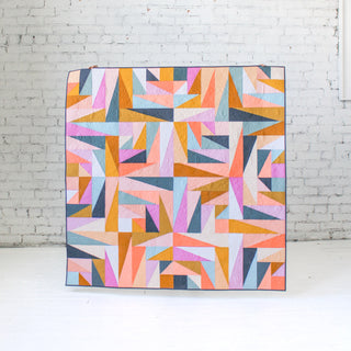 Colorful geometric quilt with muted pastel shades arranged in angular shapes, displayed against a white brick wall, showcasing the Super Scrappy Repair Quilt Pattern (Download) by Eudaimonia Studio.