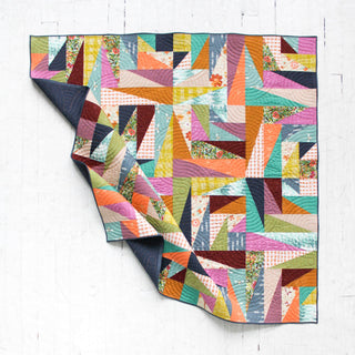 Colorful patchwork quilt with geometric shapes and a folded corner, showcasing diverse fabric patterns, paired with navy backing by Eudaimonia Studio for Super Scrappy Repair Quilt Pattern (Download)