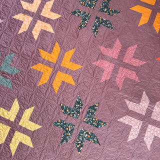 Close-up of a colorful quilt featuring star-shaped blocks with floral and solid fabrics on a purple background, showcasing the Super Star Quilt Pattern (Download) by A Piece of Quiet Quilts.