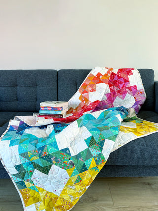 Brightly colored quilt with geometric triangle patterns in blues, yellows, pinks, and oranges draped over a dark gray sofa, showcasing the Sweet Darlings Quilt Pattern (Download) by Lou Orth Designs.