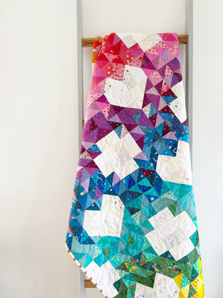 Quilt draped on a ladder featuring vibrant geometric patterns in reds, purples, blues, and greens with white accents, showcasing the Sweet Darlings Quilt Pattern (Download) by Lou Orth Designs.
