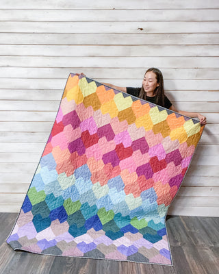 Person holding a colorful quilt with a tessellated heart pattern against a wooden background