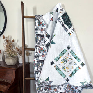 Quilt featuring floral and geometric blocks hanging on a wooden ladder beside a vintage piano, showcasing Timber & Glow Quilt Pattern (Download) by Sew & Sew Quilting Co.