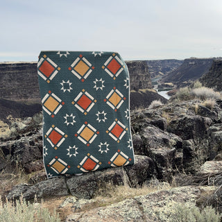 Timber & Glow Quilt Pattern by Sew & Sew Quilting Co. cover: dark teal quilt with orange and white geometric star diamond pattern propped against a rocky desert canyon landscape.