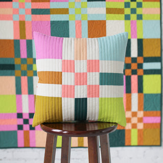 Square quilted pillow with soft pastel and earthy tones featuring geometric blocks in front of a colorful quilt, showcasing To The Nines Quilt Pattern (Download) by Patternly Collective.