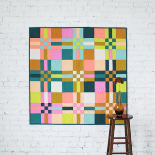 Colorful To The Nines quilt by Patternly  on a white brick wall with a wooden stool and vase.
